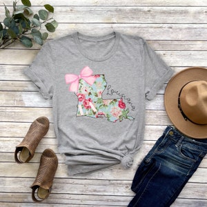 May include: Gray t-shirt with a floral print of the state of Louisiana and a pink bow. The text "Louisiana" is written in cursive on the shirt.