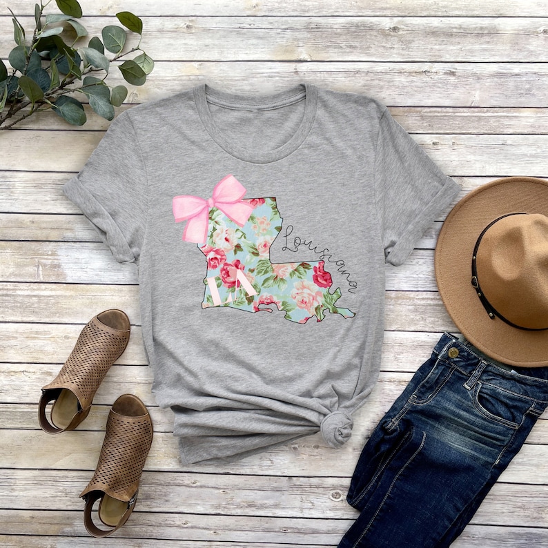 May include: Gray t-shirt with a floral print of the state of Louisiana and a pink bow. The text "Louisiana" is written in cursive on the shirt.