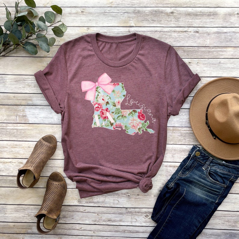 May include: A maroon t-shirt with a floral print of the state of Louisiana and a pink bow. The shirt is laid out on a wooden surface with a pair of brown sandals, a pair of blue jeans, and a brown hat.