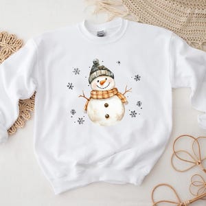 May include: White sweatshirt featuring a watercolor snowman design. The snowman wears a gray and black hat, an orange and tan scarf, and has a carrot nose. Black snowflakes are scattered around the snowman, creating a winter theme.