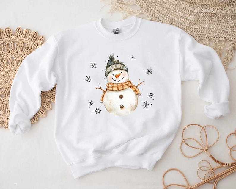 May include: White sweatshirt featuring a watercolor snowman design. The snowman wears a gray and black hat, an orange and tan scarf, and has a carrot nose. Black snowflakes are scattered around the snowman, creating a winter theme.