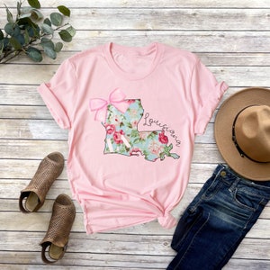 May include: Pink t-shirt with a floral print of the state of Louisiana and a pink bow. The text "Louisiana" is written in cursive.