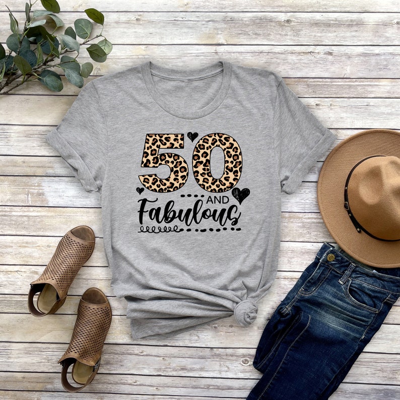 Puede incluir: Camiseta gris con un 50 estampado de leopardo y el texto "and Fabulous" en negro.