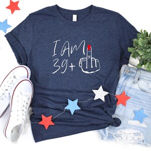 May include: A navy blue t-shirt with white text that reads "I am 39+" and a white hand with a red fingernail giving the middle finger. The shirt is laid out on a white surface with red, white, and blue stars and a pair of white sneakers.