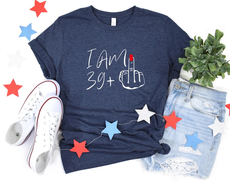 May include: A navy blue t-shirt with white text that reads "I am 39+" and a white hand with a red fingernail giving the middle finger. The shirt is laid out on a white surface with red, white, and blue stars and a pair of white sneakers.