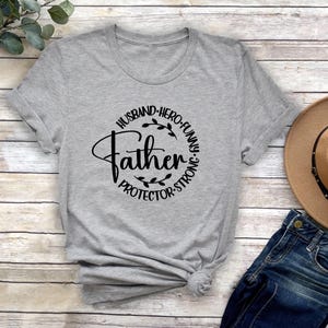 May include: Grey t-shirt with a black circle design that says "Husband, Hero, Funny, Father, Protector, Strong".