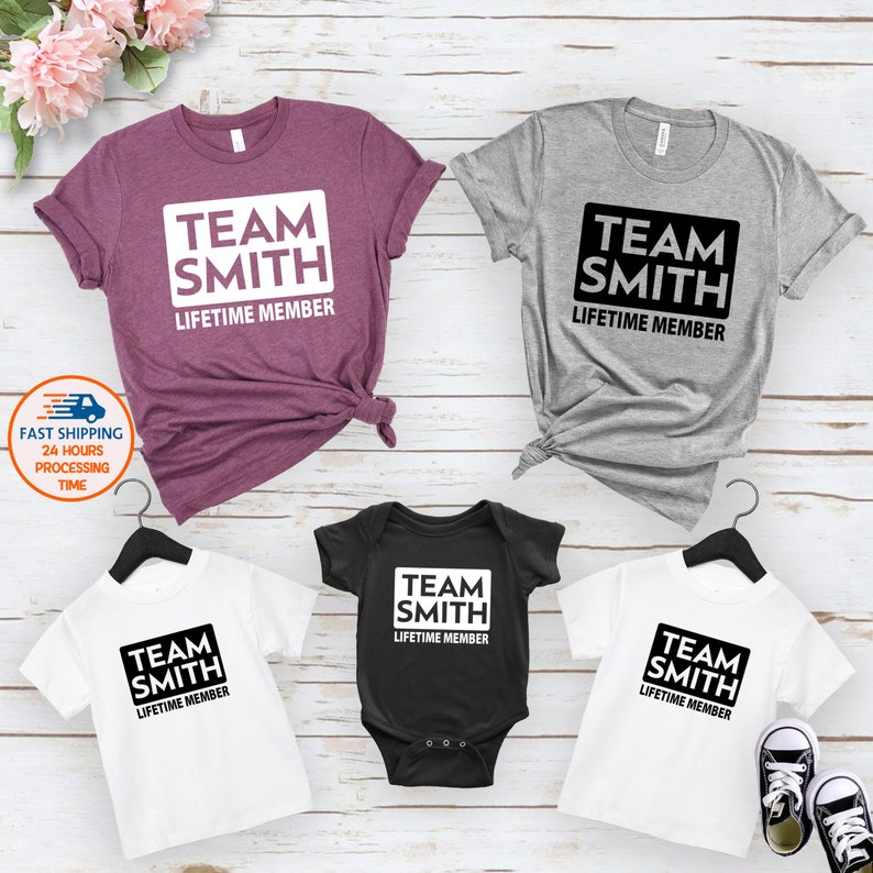 May include: A set of four t-shirts with the text "Team Smith Lifetime Member" in a black rectangle. The shirts are in different colors: purple, gray, black, and white. The shirts are arranged in a square shape.