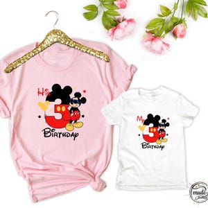 Op de afbeelding: Roze en witte T-shirts met een Mickey Mouse-afbeelding. Het roze shirt zegt "His 3rd Birthday" en het witte shirt zegt "My 3rd Birthday". Beide shirts hebben Mickey Mouse met een zonnebril en het rode nummer 3.