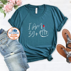 May include: Teal blue t-shirt with white text that says "I am 39+" and a white hand giving the middle finger with a red fingernail.