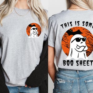 May include: A light gray t-shirt featuring a Halloween-themed design. The front has a circular graphic of a ghost wearing sunglasses. The back of the shirt has the text "THIS IS SOME BOO SHEET" with the same ghost graphic.