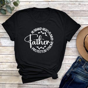 May include: Black t-shirt with a white circle graphic that says "Husband, Hero, Funny, Father, Protector, Strong".