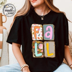 May include: Black t-shirt featuring the word "FALL" in colourful, square designs. The letters are decorated with autumn-themed patterns, including leaves and floral prints. The shirt has short sleeves and a round neckline.