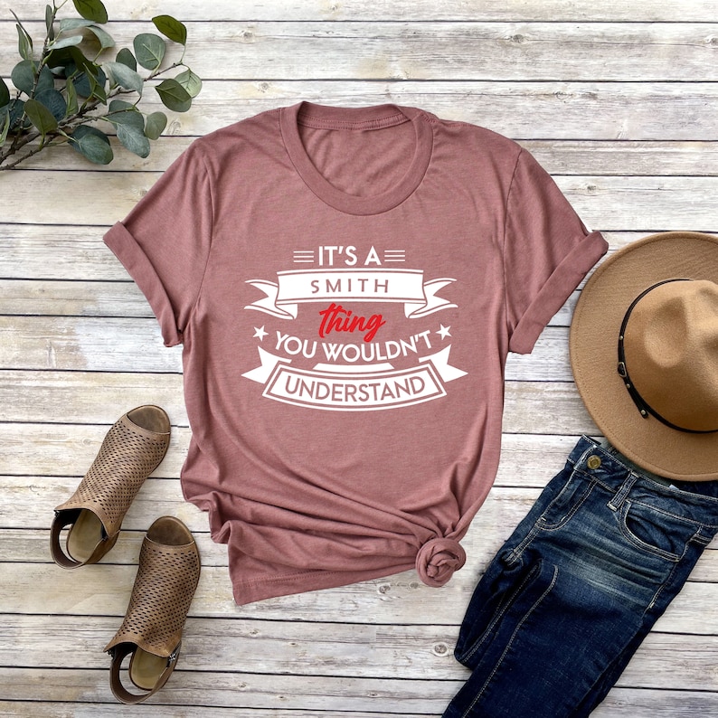 K&ouml;nnte beinhalten: Ein mauvefarbenes T-Shirt mit wei&szlig;em Text, der "It's a Smith thing you wouldn't understand" lautet. Der Text befindet sich auf einem wei&szlig;en Banner mit einem roten Stern auf jeder Seite.