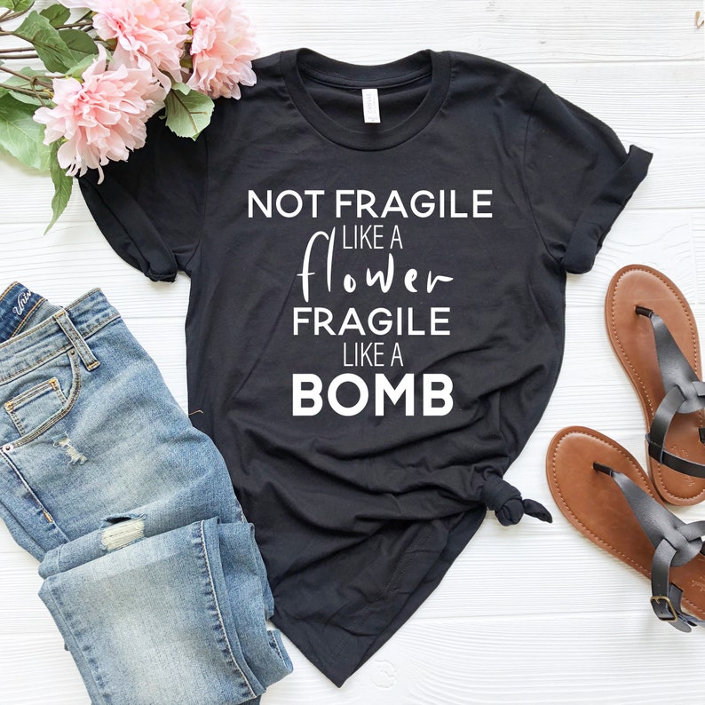 May include: Black t-shirt with the text "NOT FRAGILE LIKE A FLOWER FRAGILE LIKE A BOMB" in white. The shirt is paired with blue jeans, pink flowers, and brown sandals. The shirt is knotted at the waist.