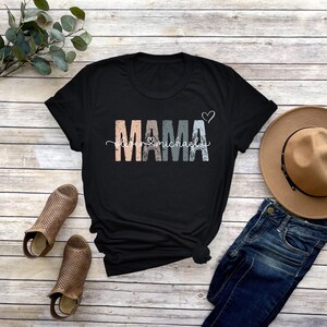May include: Black t-shirt with the word "MAMA" in colorful letters with the words "love" and "michael" underneath. The shirt is on a wooden surface with a pair of brown sandals, a pair of blue jeans, and a brown hat.
