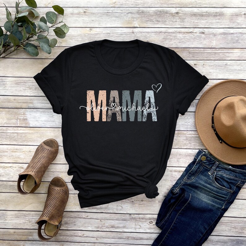 May include: Black t-shirt with the word "MAMA" in colorful letters with the words "love" and "michael" underneath. The shirt is on a wooden surface with a pair of brown sandals, a pair of blue jeans, and a brown hat.