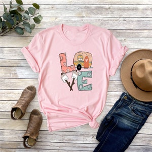 May include: Pink t-shirt with a graphic design of the word "LOVE" with a camper van and marshmallows. The letters are stylized and have a vintage look.