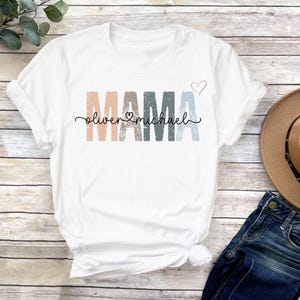 May include: White t-shirt with the word "MAMA" in colorful, distressed letters. The names "Oliver" and "Michael" are written in cursive below the word "MAMA" with a heart symbol between the names.