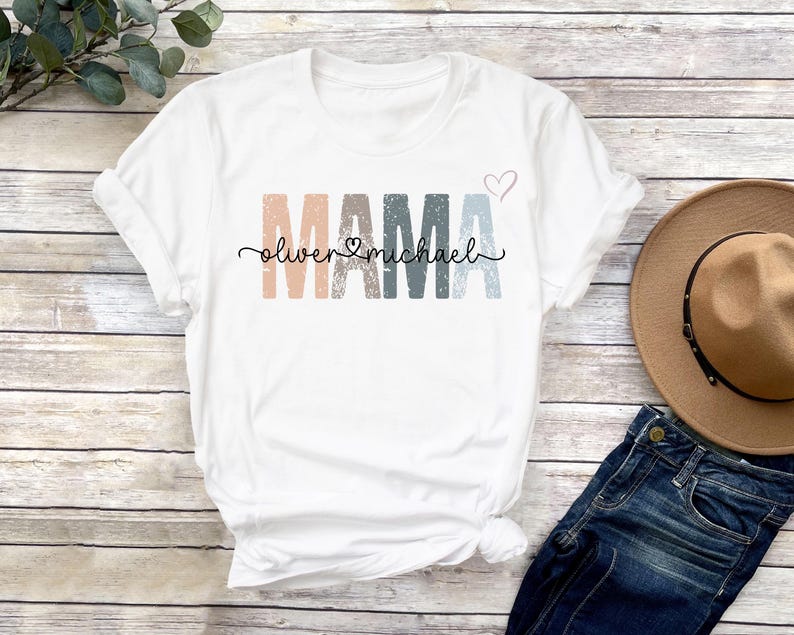 May include: White t-shirt with the word "MAMA" in colorful, distressed letters. The names "Oliver" and "Michael" are written in cursive below the word "MAMA" with a heart symbol between the names.