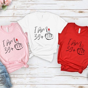May include: Three t-shirts with the text "I am 39+" and a middle finger emoji. The shirts are pink, white, and red.