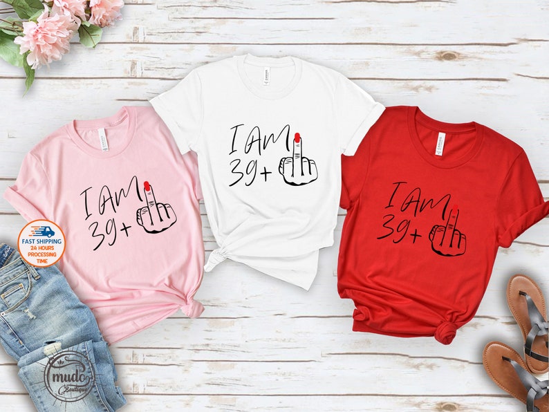 May include: Three t-shirts with the text "I am 39+" and a middle finger emoji. The shirts are pink, white, and red.