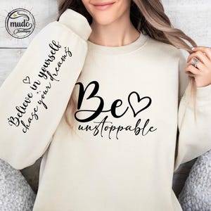 May include: Cream-colored sweatshirt with the words "Be unstoppable" in black script, with a heart symbol. The sleeve displays the text "Believe in yourself, chase your dreams". A small logo is visible in the upper left corner.