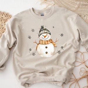 May include: A beige sweatshirt featuring a watercolor snowman design. The snowman wears a gray hat, an orange and brown scarf, and has a carrot nose. Black snowflakes are scattered around the snowman, creating a winter theme.