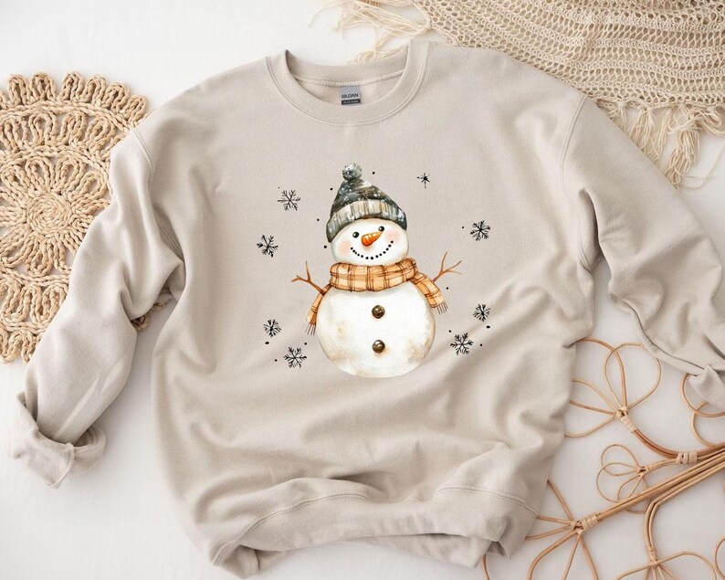 May include: A beige sweatshirt featuring a watercolor snowman design. The snowman wears a gray hat, an orange and brown scarf, and has a carrot nose. Black snowflakes are scattered around the snowman, creating a winter theme.