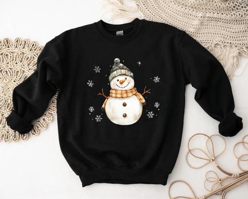 May include: Black sweatshirt featuring a watercolor snowman design. The snowman wears a gray and white knit hat, an orange and tan plaid scarf, and has twig arms. White snowflakes are scattered around the snowman. A cozy winter-themed apparel item.
