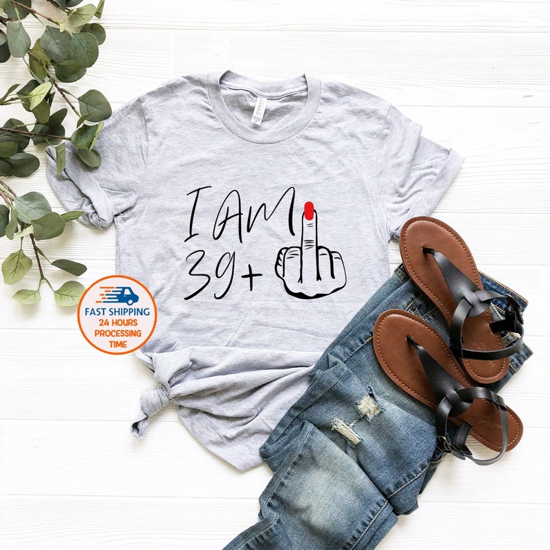 May include: Gray t-shirt with the text "I Am 39+" and a black and white illustration of a middle finger with red nail polish.