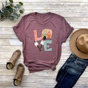 May include: A maroon t-shirt with a graphic of the word "LOVE" with a camper van and marshmallows. The letters are made of different patterns and colors.