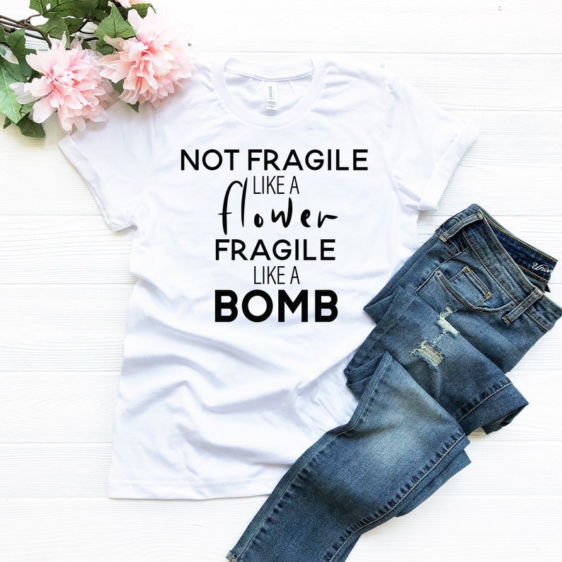 May include: White t-shirt with the text "NOT FRAGILE LIKE A FLOWER FRAGILE LIKE A BOMB" in black. The shirt is paired with blue denim jeans and pink flowers.