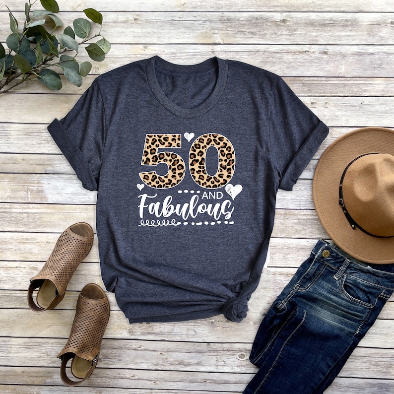 Puede incluir: Una camiseta gris oscuro con un dise&ntilde;o de estampado de leopardo del n&uacute;mero 50 y el texto "and Fabulous" en blanco. La camiseta est&aacute; colocada sobre una superficie de madera con un par de sandalias marrones, un sombrero marr&oacute;n y unos vaqueros azules.