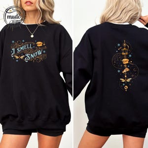 May include: Black sweatshirt with the text "I Smell Snow" in teal and gold script, featuring dragonfly and snowflake designs. The back of the sweatshirt has a matching design. A cozy winter-themed apparel item.