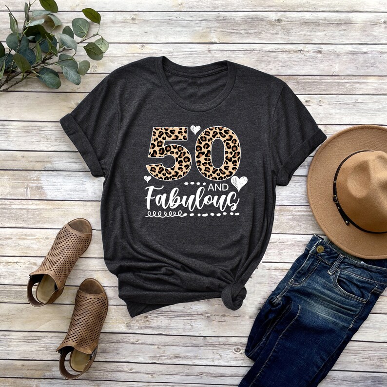 Puede incluir: Una camiseta gris oscuro con un estampado de leopardo 50 y el texto "AND Fabulous" en blanco. La camiseta est&aacute; colocada sobre una superficie de madera con un par de sandalias marrones, un sombrero marr&oacute;n y unos vaqueros azules.
