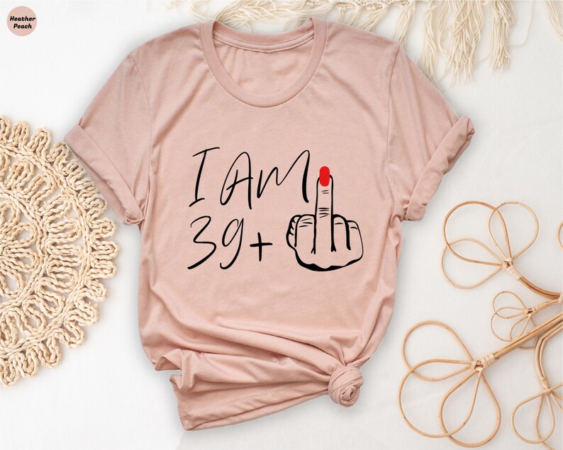 May include: A light pink t-shirt with the text "I Am 39 +" and a black and red illustration of a middle finger.