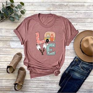 May include: A mauve t-shirt with a graphic design of the word "LOVE" with a camper van inside the "O". The letters are decorated with swirls and dots. There are two marshmallows on sticks in front of the "V".