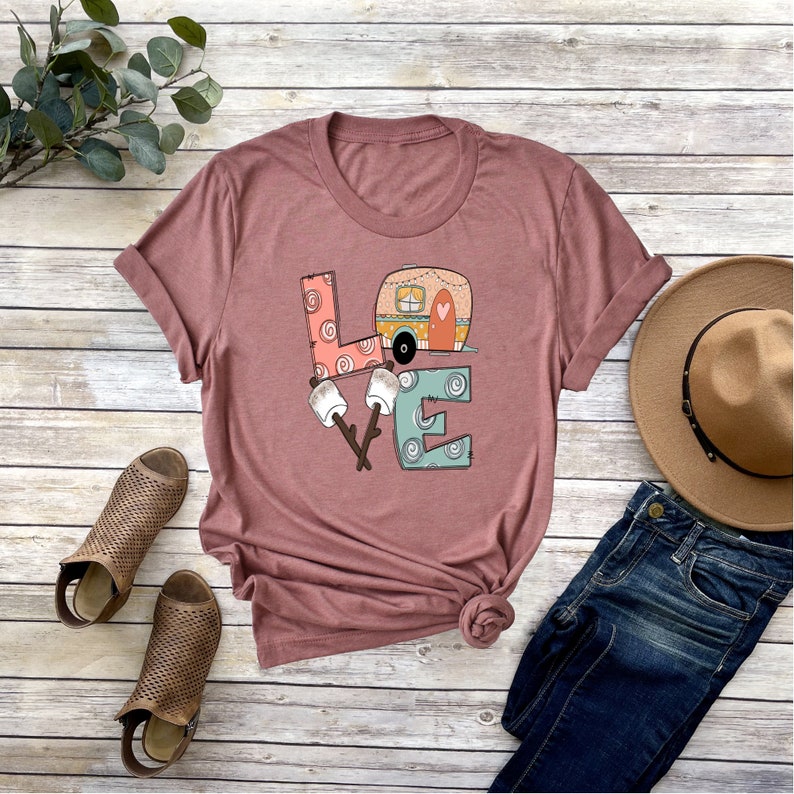 May include: A mauve t-shirt with a graphic design of the word "LOVE" with a camper van inside the "O". The letters are decorated with swirls and dots. There are two marshmallows on sticks in front of the "V".