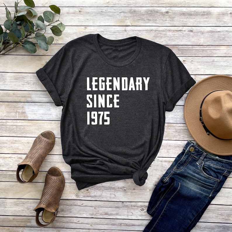 May include: A dark gray t-shirt with the text "LEGENDARY SINCE 1975" printed in white on the front. The shirt is laid flat on a wooden surface with a pair of brown sandals, a pair of blue jeans, and a brown hat.
