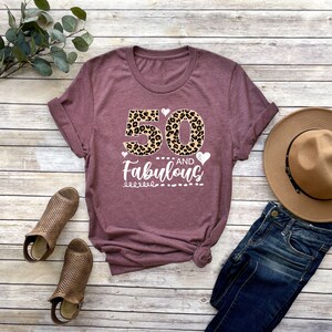 Puede incluir: Una camiseta de color marr&oacute;n con un 50 estampado de leopardo y el texto "and Fabulous" en blanco. La camiseta est&aacute; colocada sobre una superficie de madera con un par de sandalias marrones, un sombrero marr&oacute;n y unos vaqueros azules.