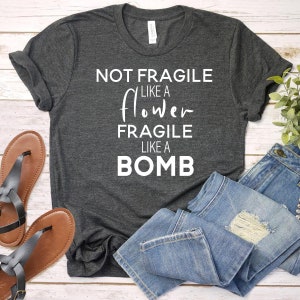 May include: Dark gray t-shirt with the text "NOT FRAGILE LIKE A FLOWER FRAGILE LIKE A BOMB" in white. The shirt is paired with blue jeans and brown sandals.
