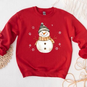May include: A red sweatshirt featuring a snowman graphic. The snowman wears a gray and white knit hat, a plaid scarf, and has a carrot nose. White snowflakes are scattered around the snowman. The sweatshirt is a long-sleeved crew neck.