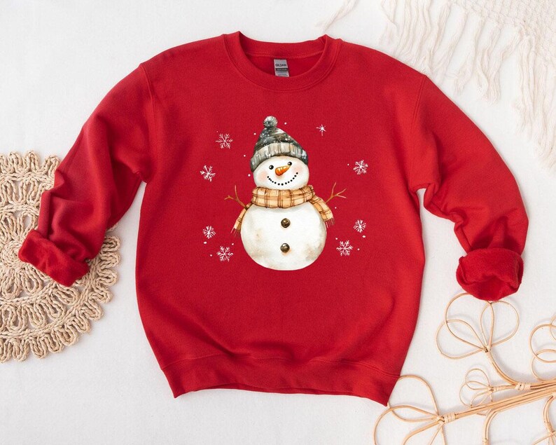 May include: A red sweatshirt featuring a snowman graphic. The snowman wears a gray and white knit hat, a plaid scarf, and has a carrot nose. White snowflakes are scattered around the snowman. The sweatshirt is a long-sleeved crew neck.