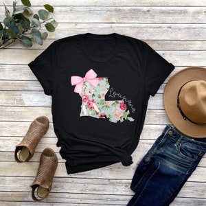 May include: Black t-shirt with a floral print of the state of Louisiana with a pink bow on top. The text "Louisiana" is written in white cursive on the right side of the state outline.