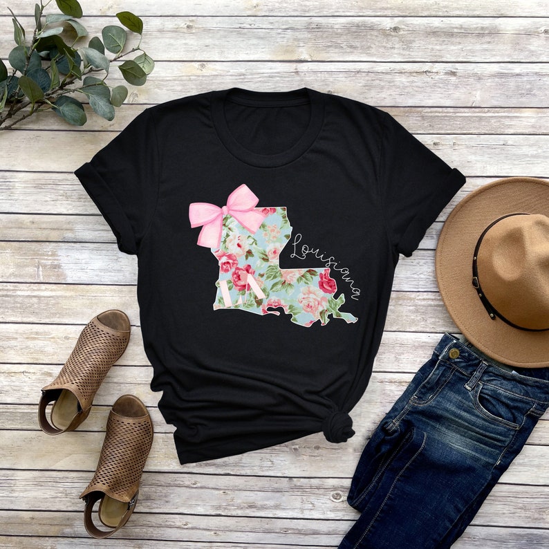 May include: Black t-shirt with a floral print of the state of Louisiana with a pink bow on top. The text "Louisiana" is written in white cursive on the right side of the state outline.
