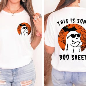 May include: White t-shirt featuring a Halloween-themed design. The front has a small circular graphic of a ghost wearing sunglasses. The back of the shirt has a larger graphic with the text "THIS IS SOME BOO SHEET".
