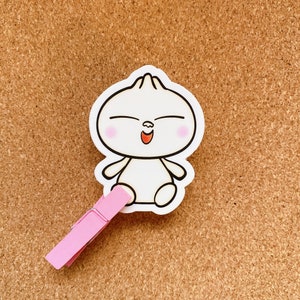 May include: A white cartoon character with a smiling face and closed eyes. The character has a round body and two small arms. The character is attached to a pink clothespin.