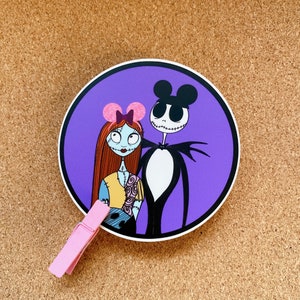 May include: A purple circle sticker with a cartoon image of Jack Skellington and Sally from the Nightmare Before Christmas. Jack is wearing a black and white suit and Sally is wearing a pink bow and a blue and yellow dress.