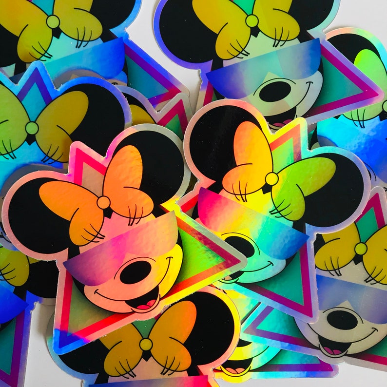 May include: Holographic stickers featuring a cartoon mouse wearing sunglasses and a bow. The stickers have a vibrant, rainbow-like color scheme with a retro geometric design. The mouse is smiling.