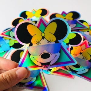 May include: Holographic sticker featuring a smiling cartoon mouse with black ears, a yellow bow, and sunglasses. The mouse is set against a teal and pink triangle. The sticker has a shiny, iridescent finish.